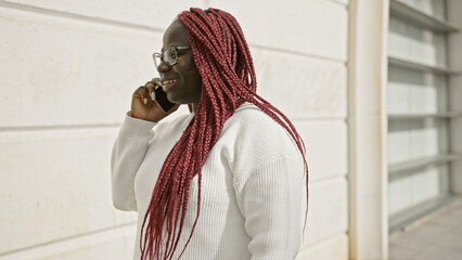 African american woman with red braids on a call, standing outdoors in an urban setting, exuding...