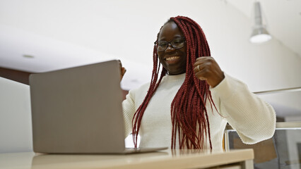 A joyful african american woman with braids working on a laptop in a modern office interior.