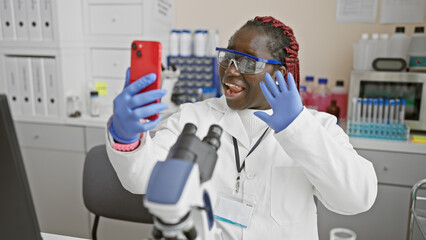 African woman with braids in a lab having a video call on her smartphone, expressing gladness.