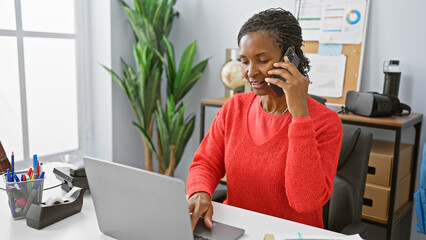 African american woman in office multitasking with smartphone and laptop, looking professional and...