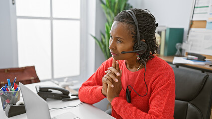 A contemplative african american woman in a headset sits at her office desk, surrounded by modern...