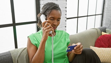 A mature african american woman talks on the phone and holds a credit card indoors.