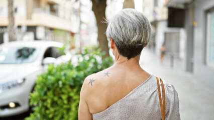 A mature woman with grey short hair and a tattoo walks in an urban setting, embodying elegance.