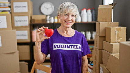A mature woman with short grey hair, wearing a volunteer shirt, holds a heart in a warehouse full...