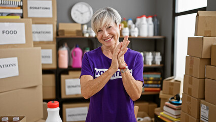 A smiling middle-aged woman with gray hair wearing a volunteer t-shirt claps in a donation center...