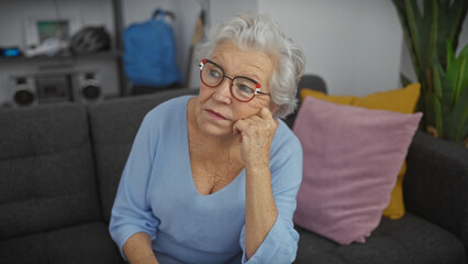 A thoughtful elderly woman with glasses sits on a sofa in a modern living room setting, expressing...