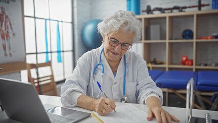A senior woman doctor writes at her desk in a clinic with rehabilitation equipment in the background