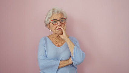 A thoughtful senior woman with gray hair stands against a pink background, portraying contemplation...