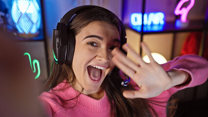 A joyful young hispanic woman wearing headphones engages in a lively video call inside a gaming room at night.