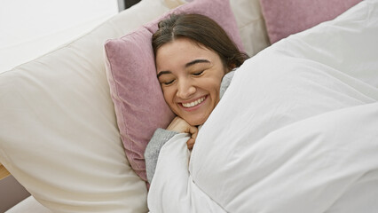 A smiling young hispanic woman enjoying relaxation in a cozy bedroom setting, exuding happiness and...