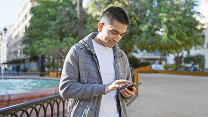 Handsome hispanic man using smartphone in a sunny urban park with trees