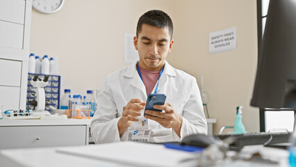 Hispanic man in white lab coat uses smartphone in laboratory setting, portraying modern healthcare...