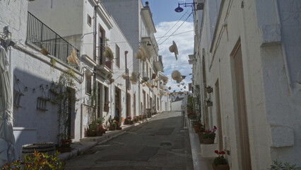Charming street in alberobello, italy, puglia, decorated with hanging hats, flowers, and potted plants under a blue sky in a historic town setting.