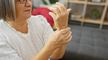 Mature woman experiences wrist pain in living room, indicative of arthritis or injury.