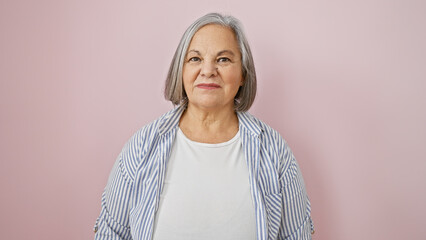 A confident senior woman with grey hair stands against a pink background, exuding elegance and...