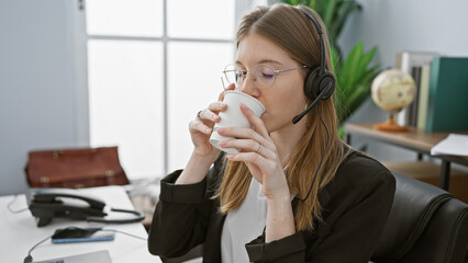 A young woman wearing a headset sips coffee in a modern office, exemplifying a professional...