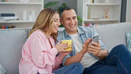 Man and woman couple drinking coffee using smartphone sitting on sofa at home