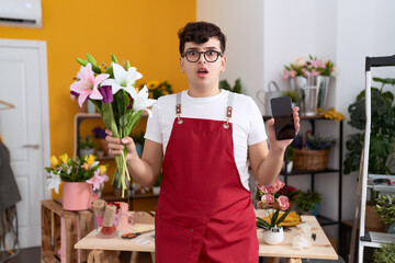 Young non binary man working at florist shop showing smartphone screen afraid and shocked with...