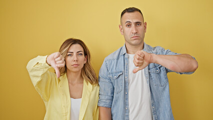 Man and woman couple standing with serious face doing thumb down gesture over isolated yellow...