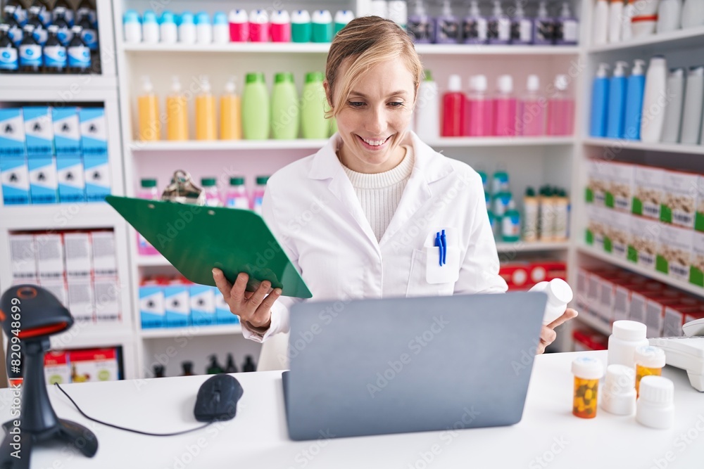 Wall mural young blonde woman pharmacist using laptop reading document at pharmacy