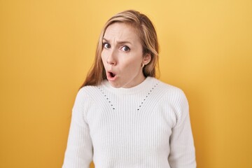 Young caucasian woman wearing white sweater over yellow background in shock face, looking skeptical...