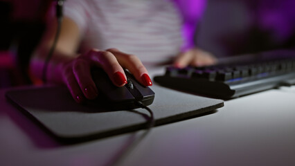 A woman with manicured nails uses a computer mouse and keyboard in a dimly lit gaming room at night.