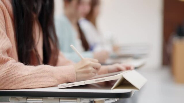 A woman is writing on a tablet with a pen. She is sitting in a classroom with other students