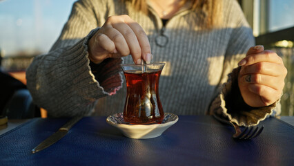 A young adult woman enjoys tea at an outdoor cafe table with a heart tattoo visible on her finger.