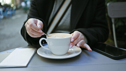 Adult woman enjoys a cappuccino at an outdoor cafe, her smartphone beside her on the table.