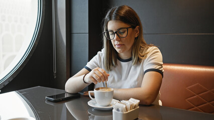 A young woman stirring coffee in a modern cafe with her smartphone on the table gives a relaxed,...