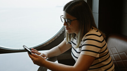 A young adult woman using a smartphone while sitting by a porthole window inside a cruise ship.