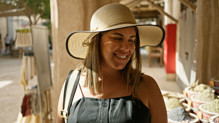 A smiling woman in a straw hat enjoys exploring a traditional market in dubai, surrounded by spices...