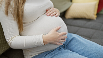 Pregnant hispanic woman caressing her belly, seated on a couch indoors with cushions in the...