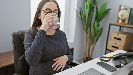 Pregnant hispanic woman drinking water while working in a modern office setting.