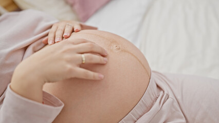 A young pregnant woman lies on a bed touching her belly in a home bedroom setting, embodying maternity and expecting.