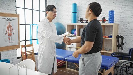 A male doctor and patient shaking hands in a physiotherapy clinic with rehabilitation equipment in...