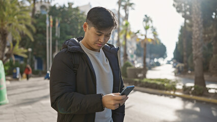 A young asian man using a smartphone stands on a sunny city street lined with palm trees.