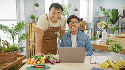 Two men smiling in a flower shop with a laptop, fresh flowers, garden tools, and indoor plants in...