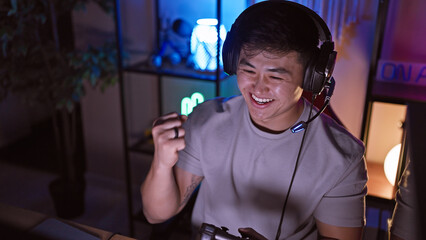 A happy asian man gaming in a dark room with led lights wearing headphones.