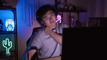 Young asian man with headphones in a dark gaming room showing signs of neck pain.