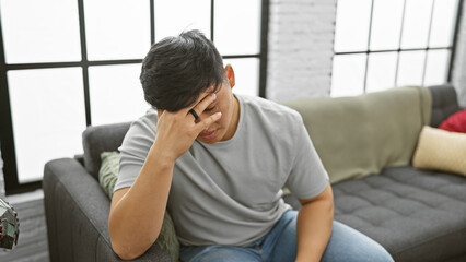 A stressed asian man sitting on a sofa indoors, covering face with hand, expressing worry or...