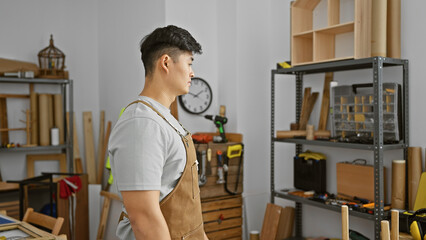 Asian man in a carpentry workshop, looking away pensively amid tools and wood.