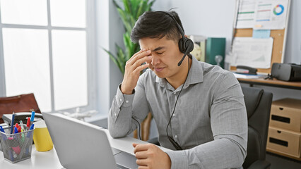 A stressed asian man with a headset in an office expressing a headache at his desk.