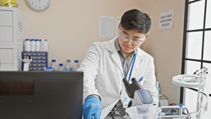 A young asian man wearing lab coat and safety glasses works with a microscope in a modern laboratory setting.