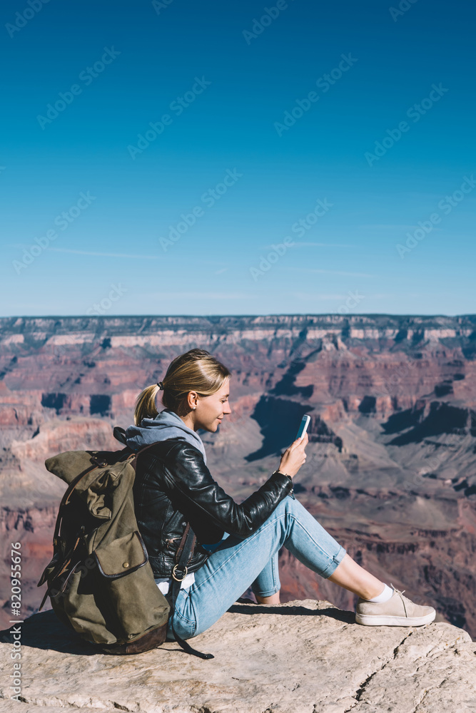 Poster smiling girl with rucksack using mobile connection for sending messages from rocky cliff in national