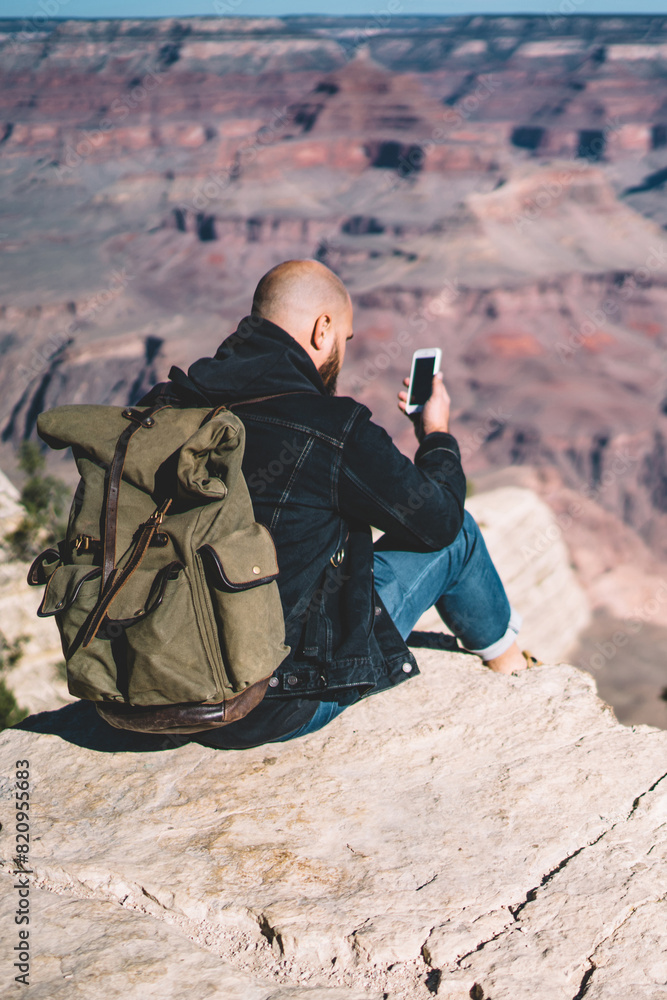 Poster hipster guy with rucksack sitting on high mountain cliff using smartphone for reading text message i