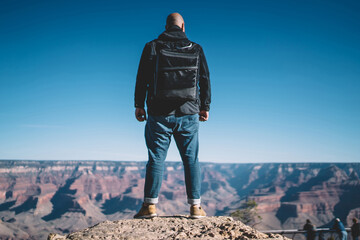 Back view of man wanderer with backpack standing on cliff of high rock observing nature...
