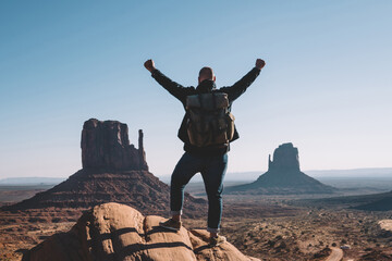 Silhouette back view of excited male traveler raising hands up standing on rock over desert with...