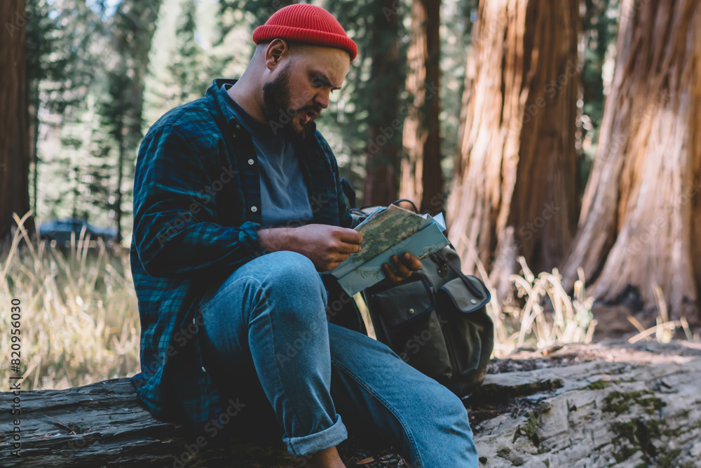 Poster concentrated hipster guy reading map checking route and direction while having rest during hiking to