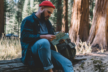 Concentrated hipster guy reading map checking route and direction while having rest during hiking...